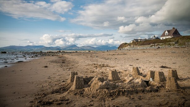 Ein sandiger Strand mit Sandburgen im Vordergrund und ein Haus mit rotem Dach im Hintergrund.
