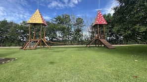 Twee houten speeltoestellen met klimnetten en glijbanen, gelegen op een grasveld met bomen op de achtergrond.