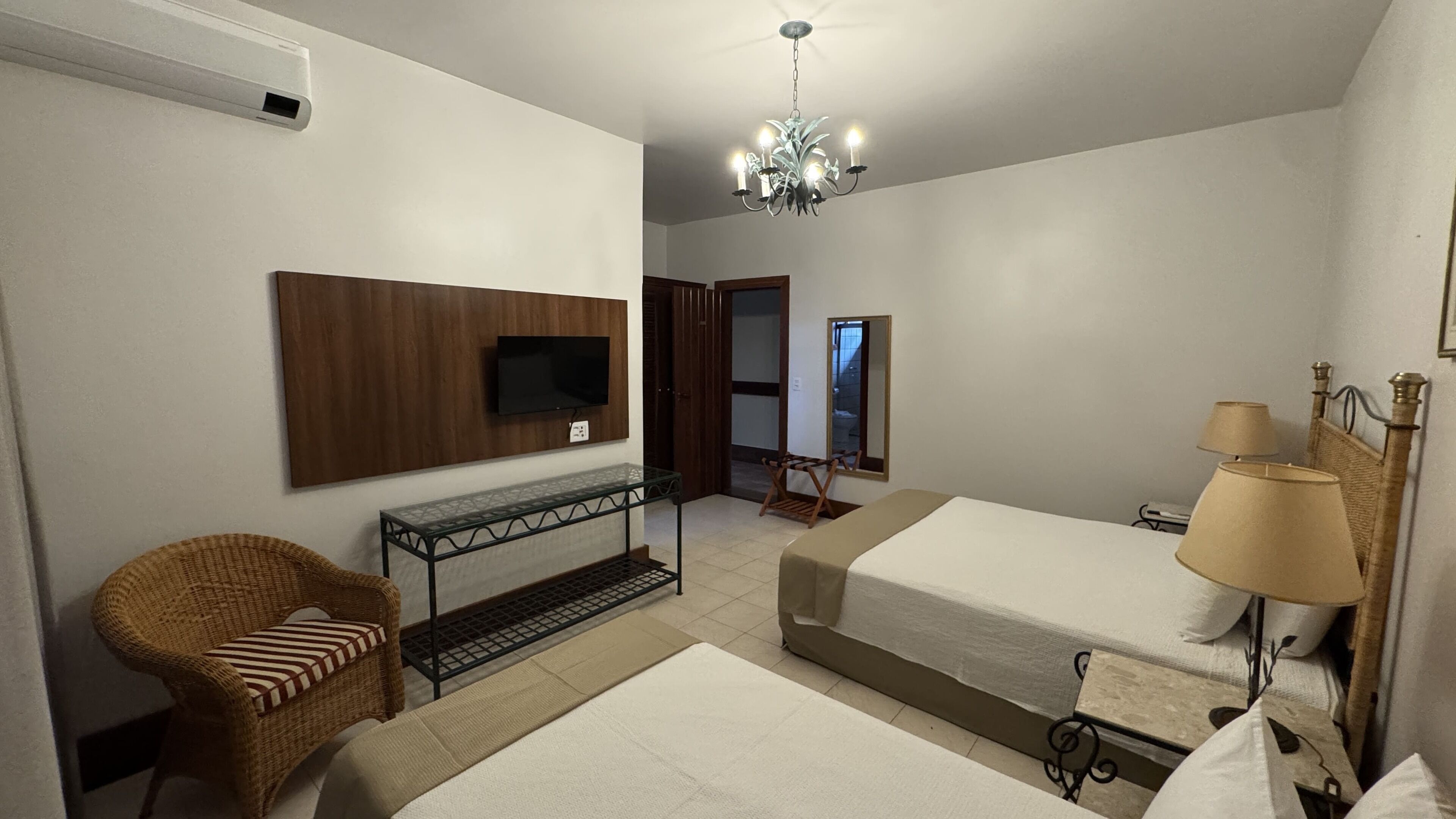 Quarto de hotel com duas camas, televisão presa na parede, cadeira de vime, mesa pequena com tampo de vidro e um lustre.