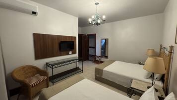 Quarto de hotel com duas camas, televisão presa na parede, cadeira de vime, mesa pequena com tampo de vidro e um lustre.