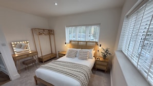 A bedroom with a large bed, a vanity with a mirror, a window with blinds, and a bedside table with a lamp. - Bader House (Ipswich)