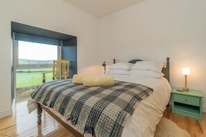 Een slaapkamer met een groot bed, een nachtkastje, een lamp en uitzicht op een groen landschap door een raam.