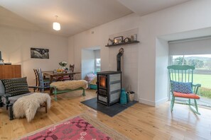 Een gezellige woonkamer met een houtkachel, een eethoek met tafel en stoelen, en uitzicht op een groen landschap door een groot raam.