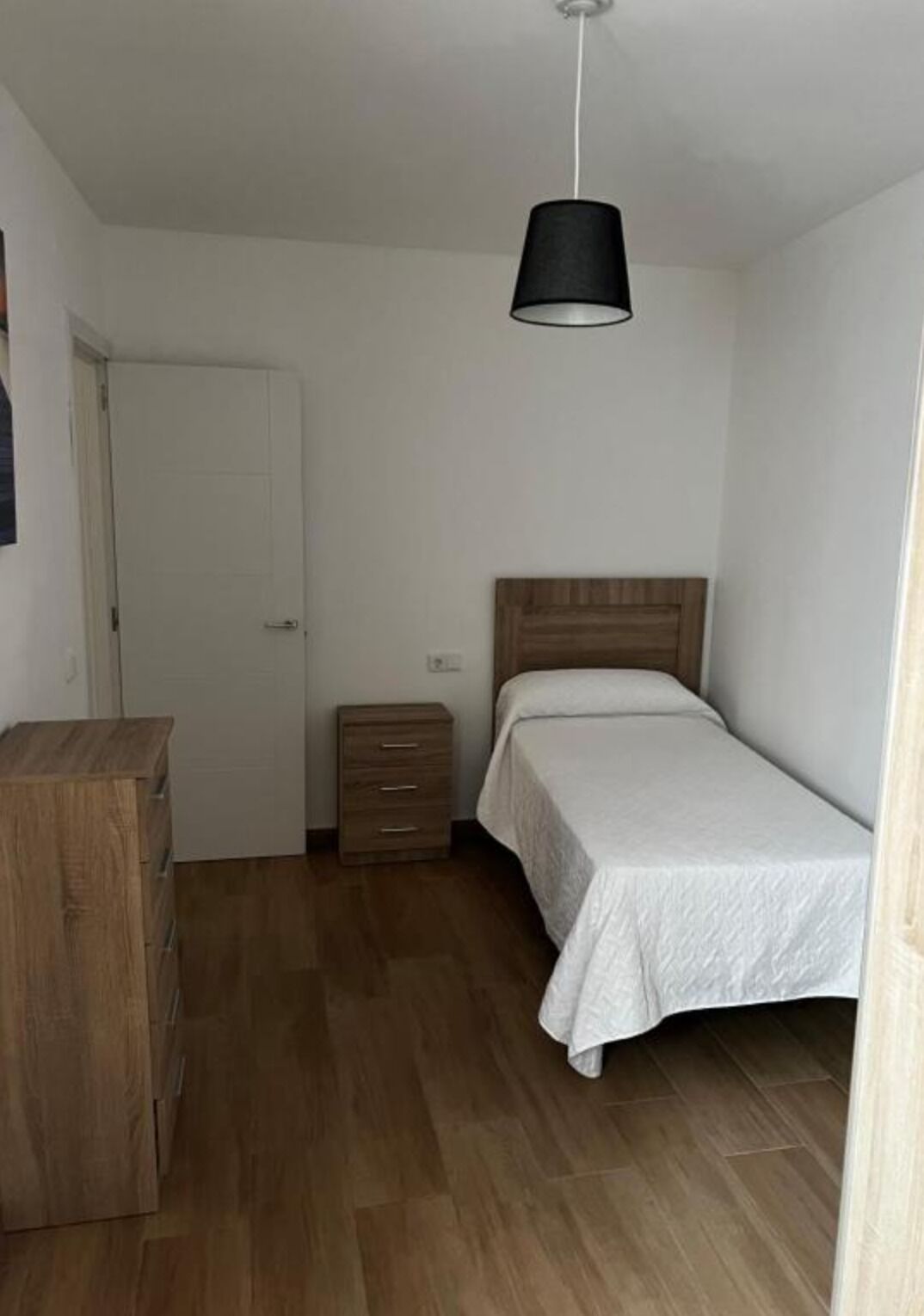 A small, clean room with a wooden dresser, a bedside table, a bed with a white bedspread, and a hanging black lamp.
