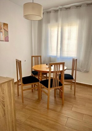 A dining area with a round wooden table, four wooden chairs with dark cushions, a hanging light fixture, and a window with sheer curtains. - Arenas Apartment (Nerja)