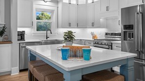 A modern kitchen with white cabinets, a large island, stainless steel appliances, and a view of the outdoors. - Private Pool + Hot Tub | New 5BR w/ Golf Cart – Steps to Beach (Emerald Isle)