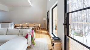 Uma sala de estar moderna com sofá, uma área de refeições com mesa e cadeiras, e uma grande janela com vista para árvores.