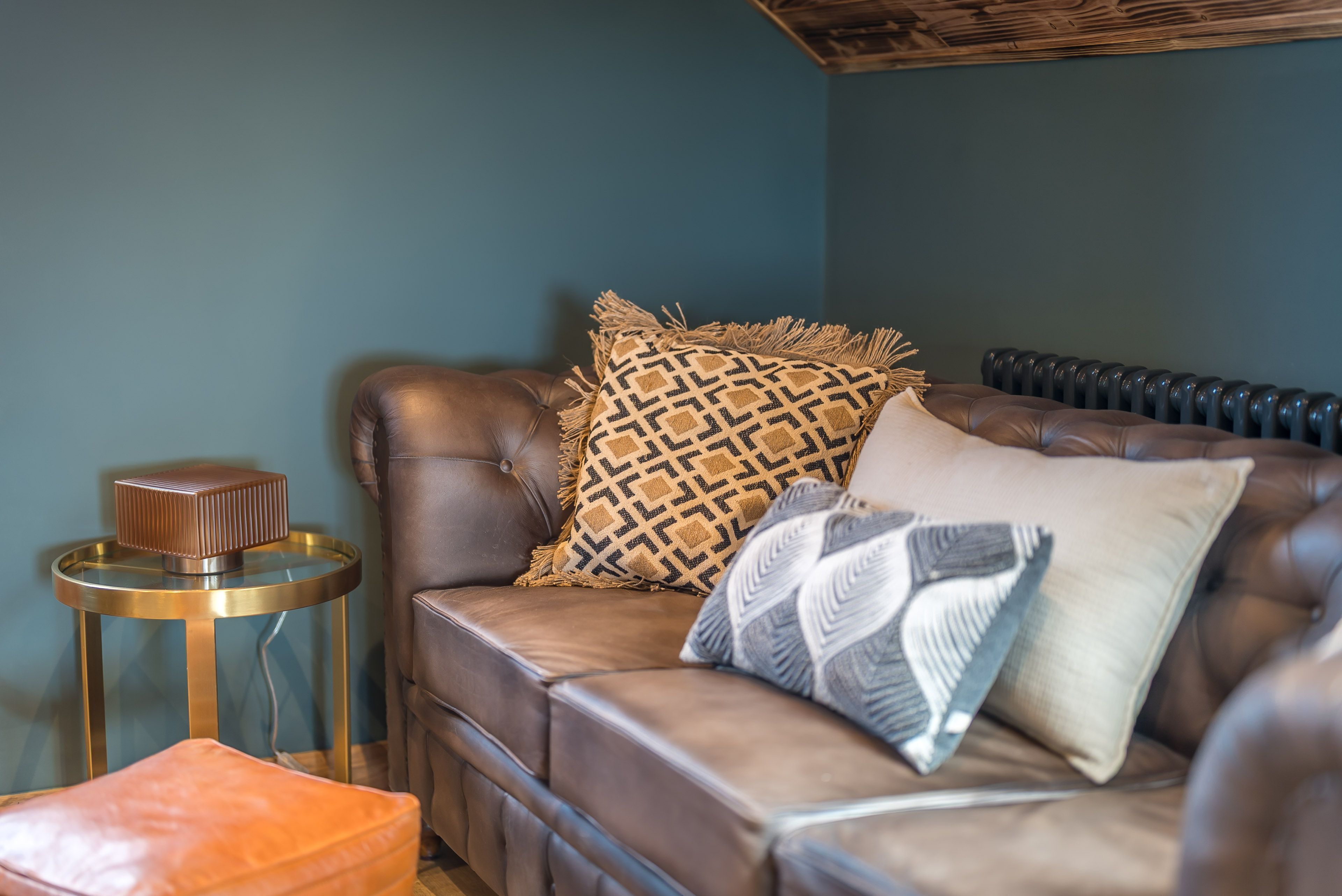 A brown leather sofa with patterned cushions, a small round side table, and a radiator against a teal wall.