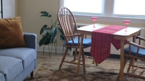 Un salón con sofá, una mesa de comedor con dos sillas, una planta en maceta y una ventana con persianas.