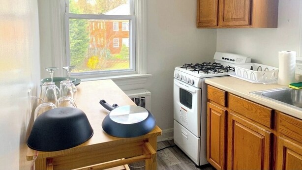 Una cocina compacta con estufa a gas, horno, muebles de madera, una ventana con persianas y una pequeña mesa de comedor con dos ollas negras.