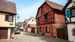 A cobblestone street lined with traditional half-timbered houses, a parked car, and a visible blue sign. - Gîte Le Nain Rouge – 18th-Century Alsatian House in Kaysersberg (Kaysersberg Vignoble)