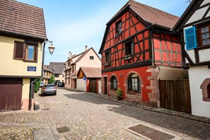 A cobblestone street lined with traditional half-timbered houses, a parked car, and a visible blue sign. - Gîte Le Nain Rouge – 18th-Century Alsatian House in Kaysersberg (Kaysersberg Vignoble)