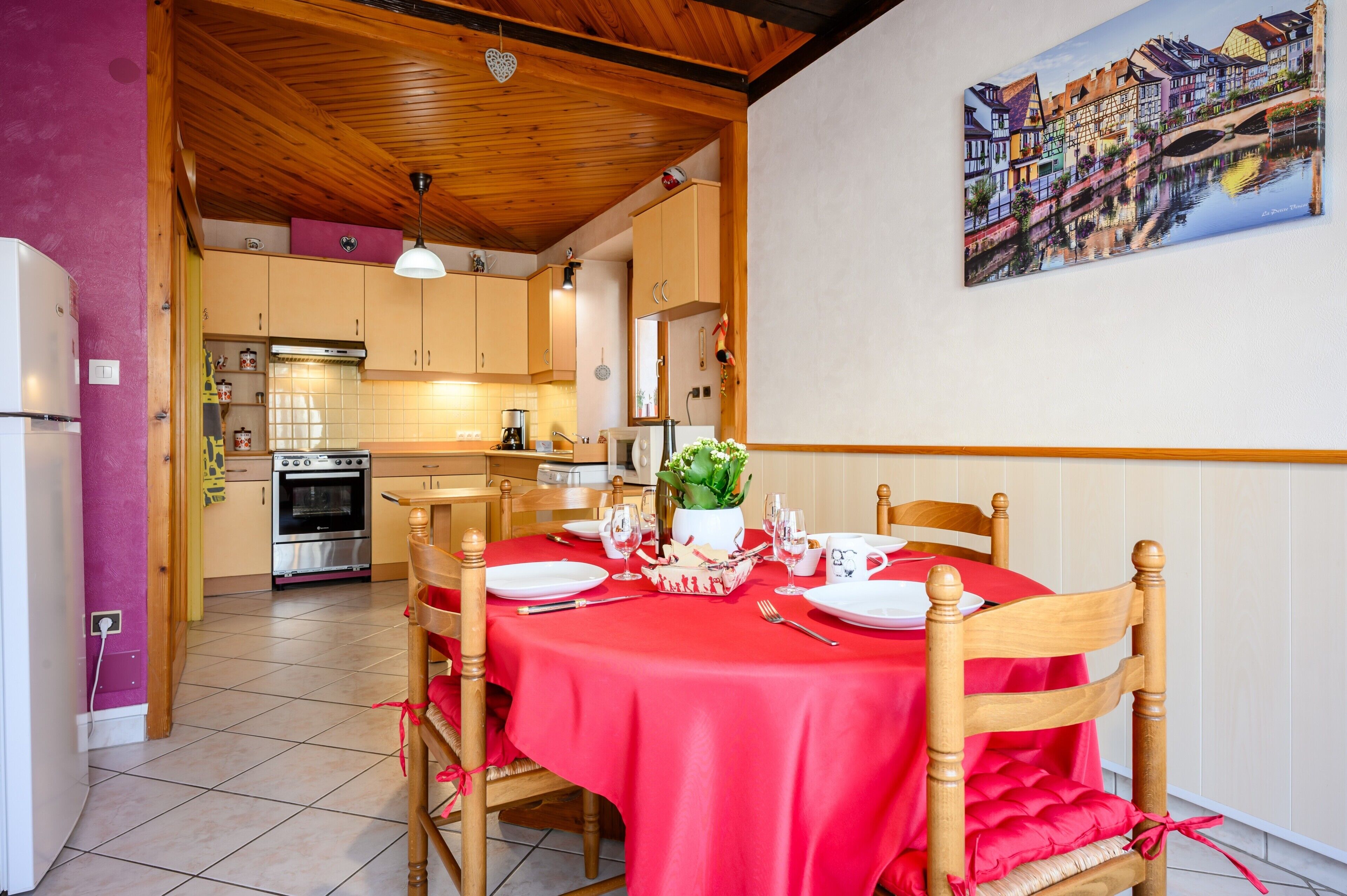 A dining area with a red tablecloth, wooden chairs, and a kitchen in the background.