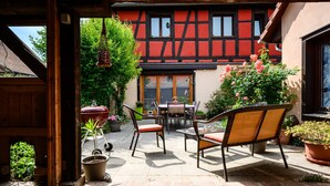 A courtyard with a table and chairs, potted plants, and a view of a traditional building with a red facade. - Gîte Le Nain Rouge – 18th-Century Alsatian House in Kaysersberg (Kaysersberg Vignoble)