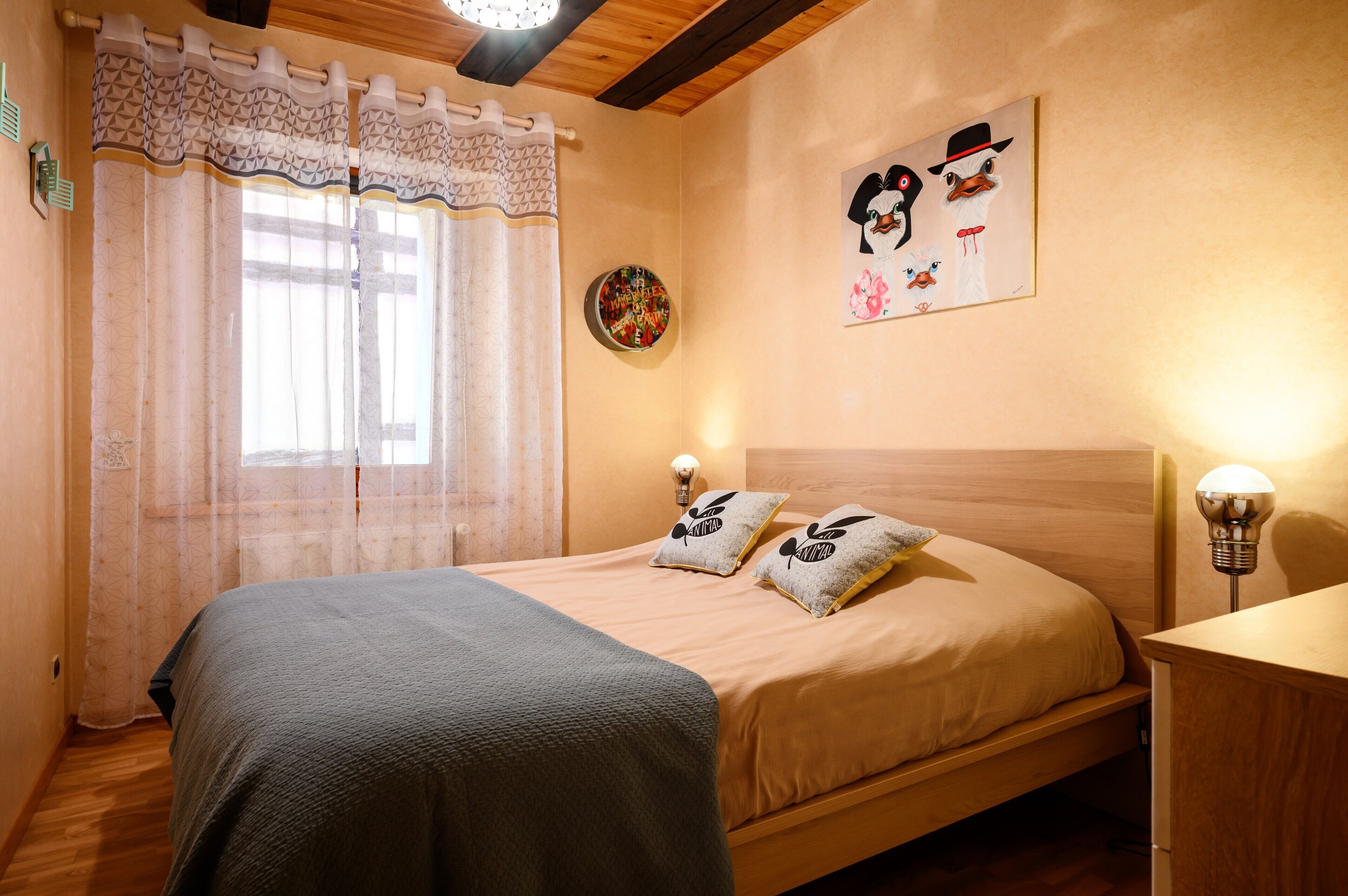 Een slaapkamer met een bed, nachtkastje, lamp, raam met gordijnen en een wanddecoratie.