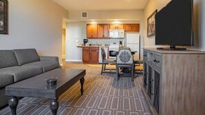 A living room with a sofa, coffee table, and a TV on a stand. There is a kitchen in the background with wooden cabinets and a microwave. - Family Friendly Condo - No Resort fees! (Las Vegas)
