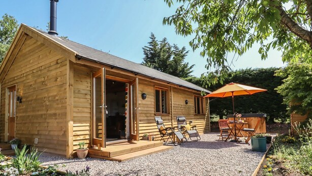 Une cabane en bois avec une porte ouverte, un patio avec des meubles d’extérieur et un jardin agrémenté de parterres de fleurs.