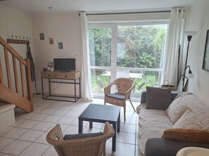 Een woonkamer met een bank, twee rieten fauteuils, een kleine tafel, een televisie op een houten dressoir en een groot raam met gordijnen.