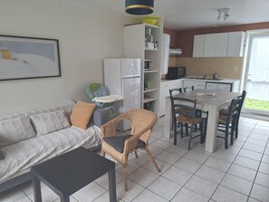Een compacte leefruimte met een bank, een eettafel, rieten stoelen, een kinderstoel, een koelkast, een magnetron en een spoelbak in de keuken.