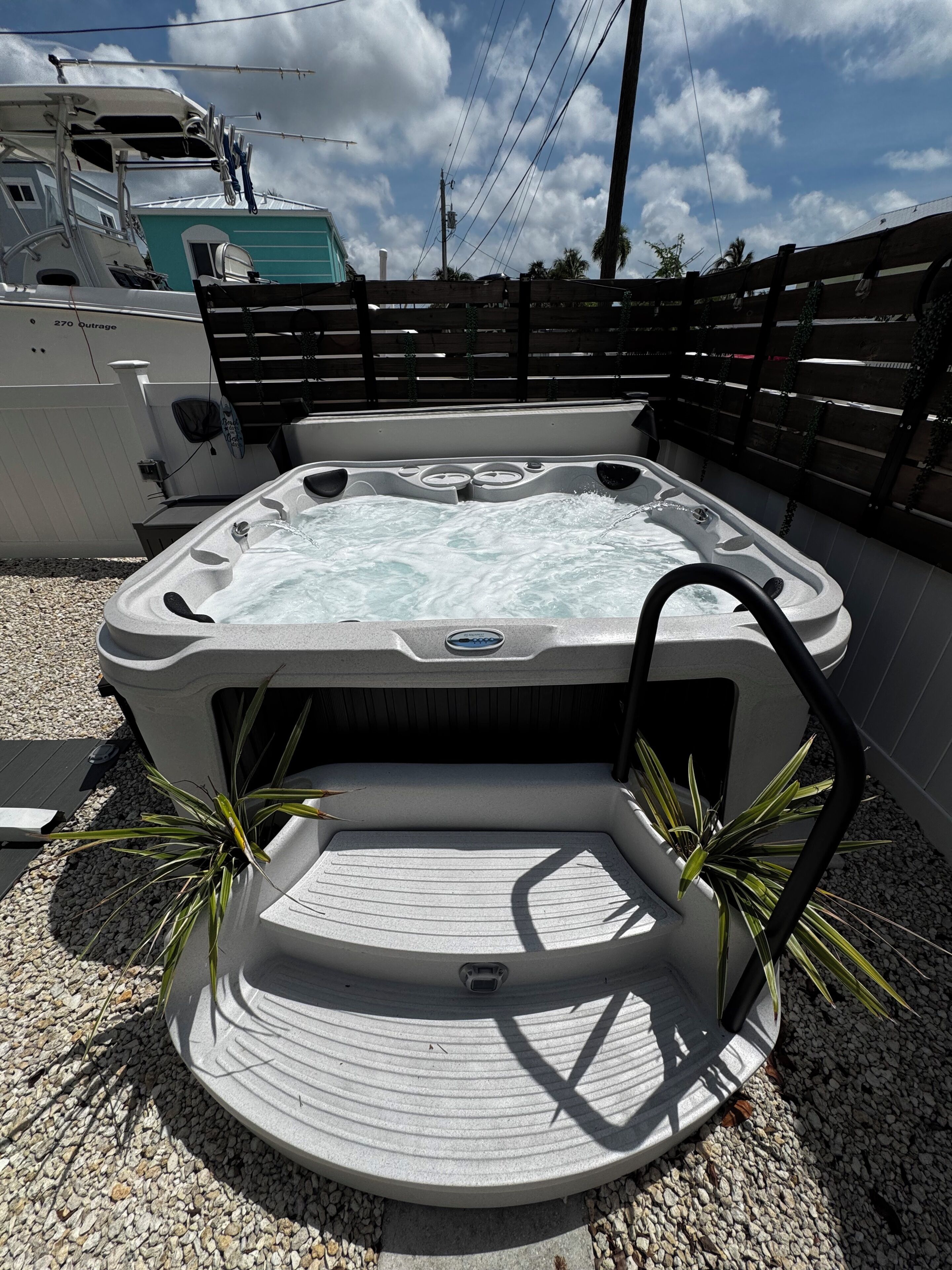 A hot tub with a control panel, surrounded by a wooden deck and gravel, with a boat and a fence in the background.