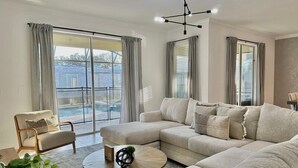 A spacious living room with a large sectional sofa, a round coffee table, and a view of a swimming pool through large sliding glass doors. - Luxury 6BR Pool & Spa Home | Near Disney 374 (Kissimmee)