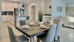 A modern kitchen with white cabinets, a central island, and a dining area with a table set for six. - Luxury 6BR Pool & Spa Home | Near Disney 374 (Kissimmee)