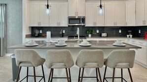 A modern kitchen with white cabinets, a central island with bar stools, a microwave, and a toaster. - Luxury 6BR Pool & Spa Home | Near Disney 374 (Kissimmee)