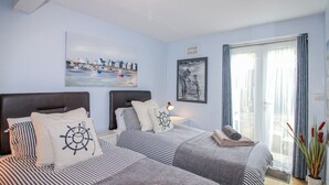 A hotel room with two beds, nautical-themed bedding, a painting of a harbor, a framed picture, and a view of the outdoors through a large window. - Quay Sands, Hope Cove (Kingsbridge)