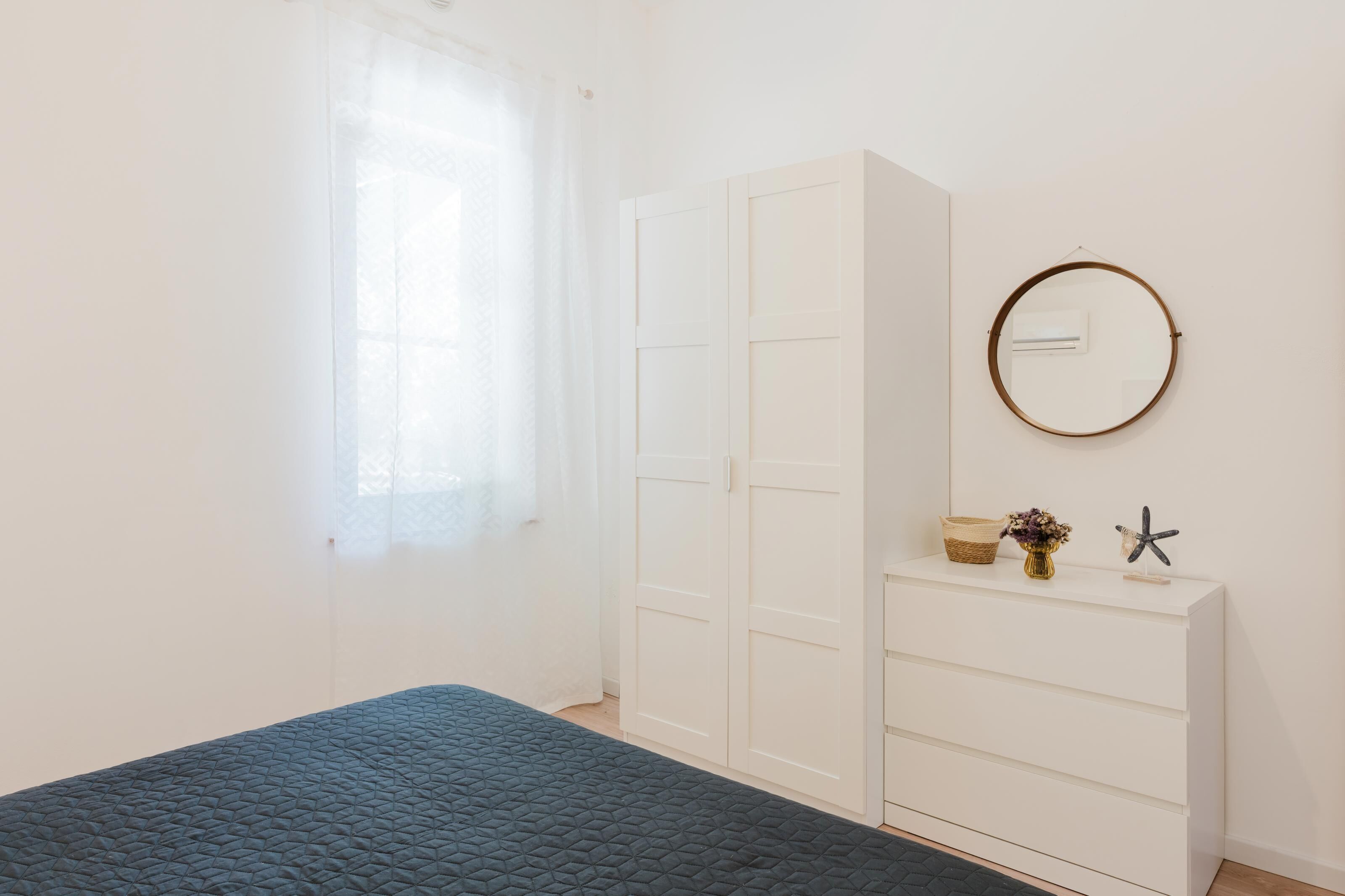 A bedroom with a bed, a wardrobe, a dresser, a window with sheer curtains, and a wall-mounted air conditioning unit.