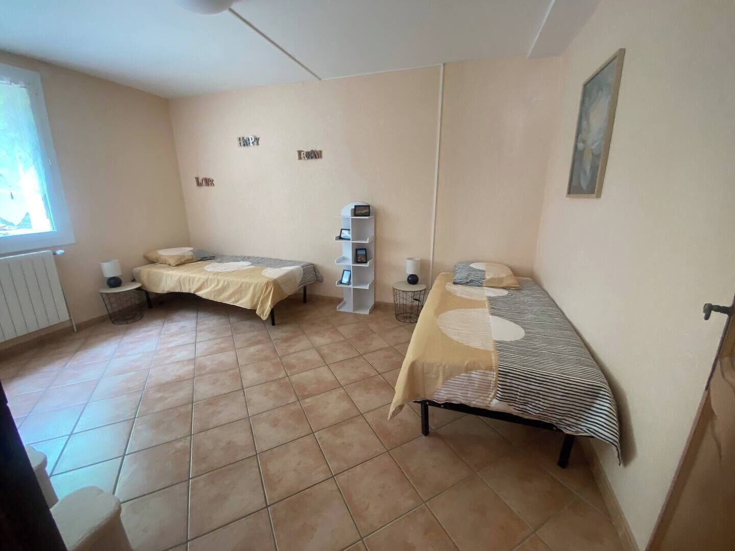 Un dormitorio pequeño y bien iluminado con dos camas, un radiador, una estantería y una ventana.