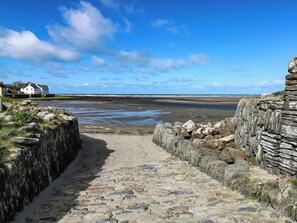 Ein Steinweg führt zu einem Strand mit Blick auf das Meer und Häuser im Hintergrund.