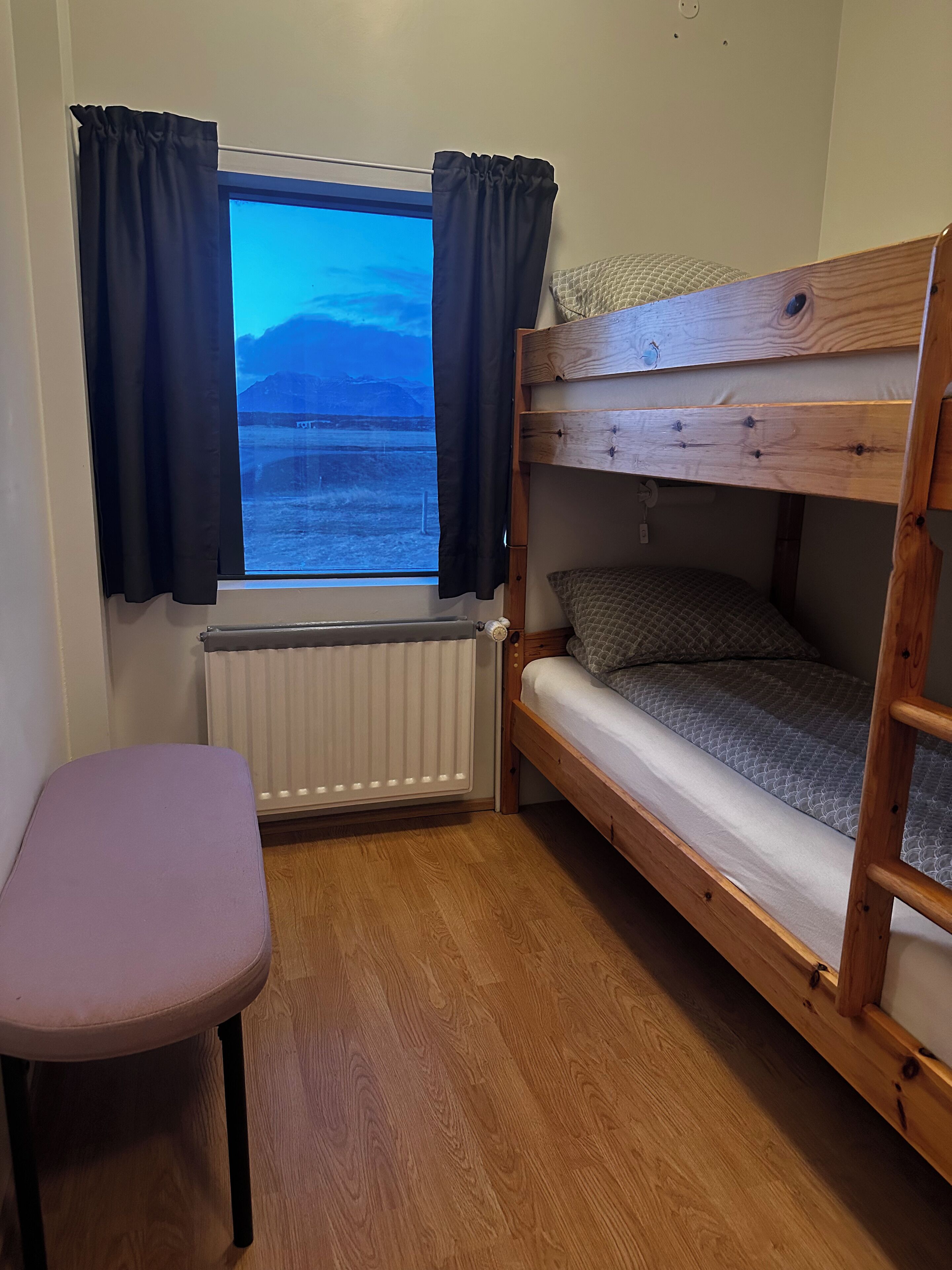 A small, cozy room with a bunk bed, a window with curtains, a radiator, and a small table.