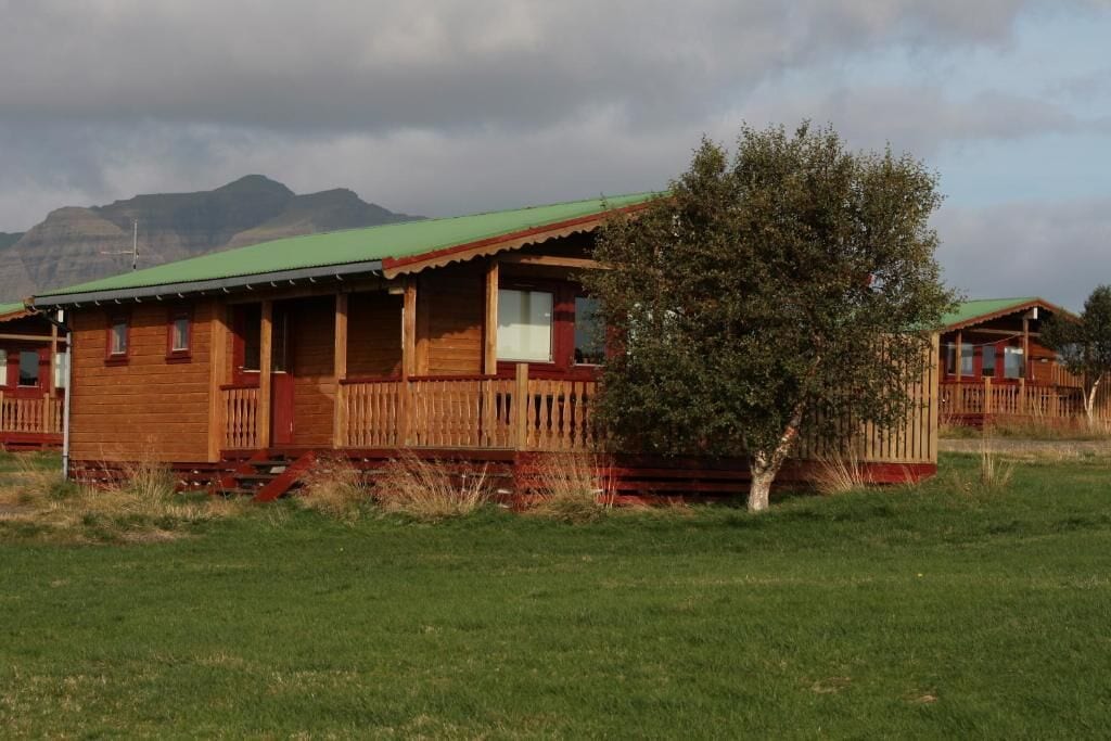 Una cabaña de madera con techo verde, rodeada por un campo cubierto de hierba y un árbol.