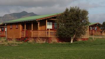 Una cabaña de madera con techo verde, rodeada por un campo cubierto de hierba y un árbol.