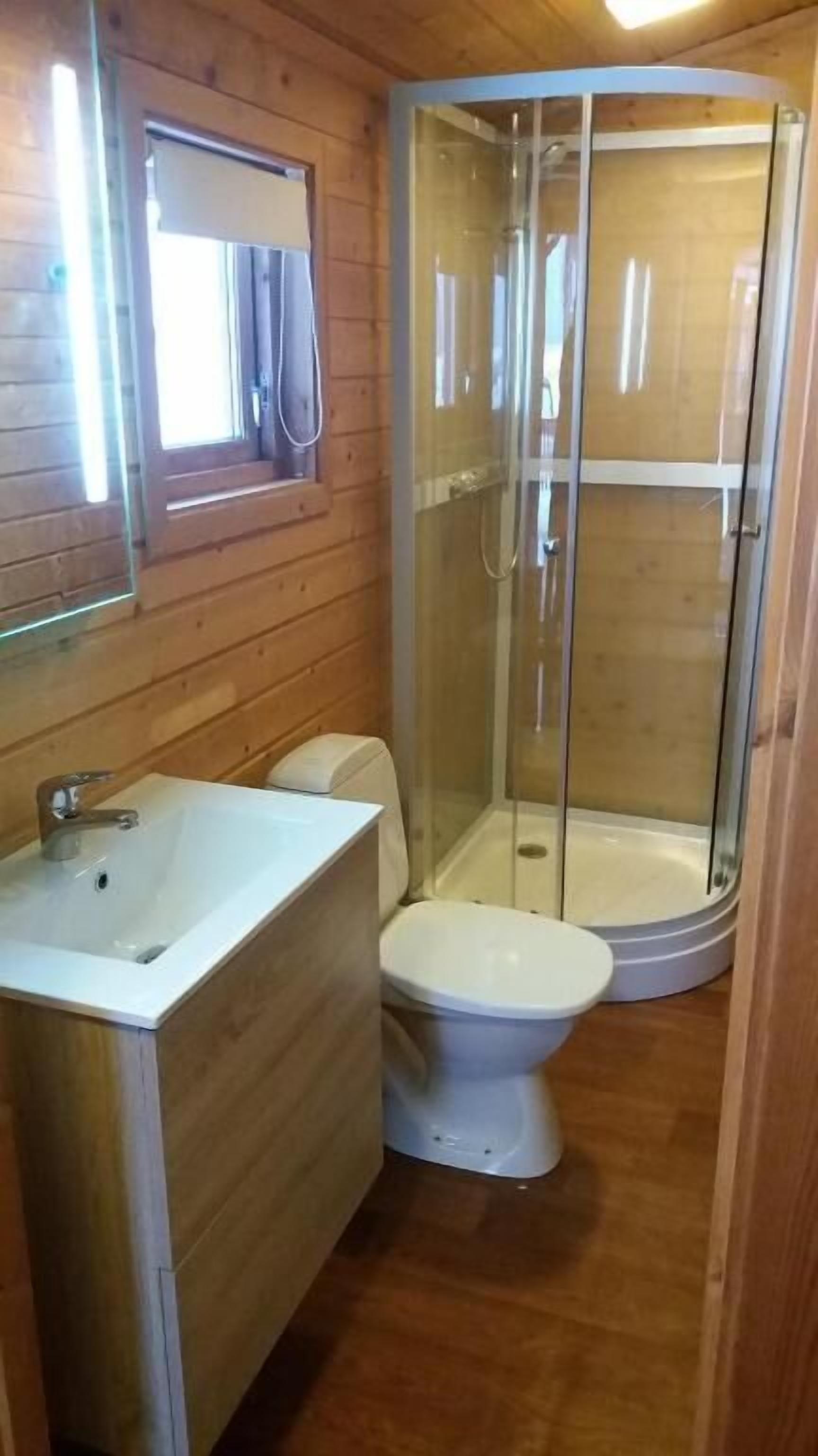 Baño de cabaña de madera con inodoro, lavamanos y ducha con cerramiento de vidrio.