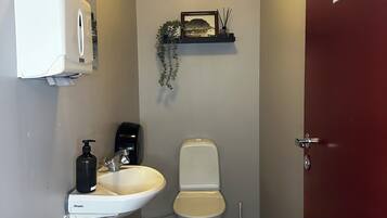 Ein Badezimmer mit Toilette, Waschbecken, Handtrockner und einem kleinen Regal mit Bilderrahmen und Pflanzen.