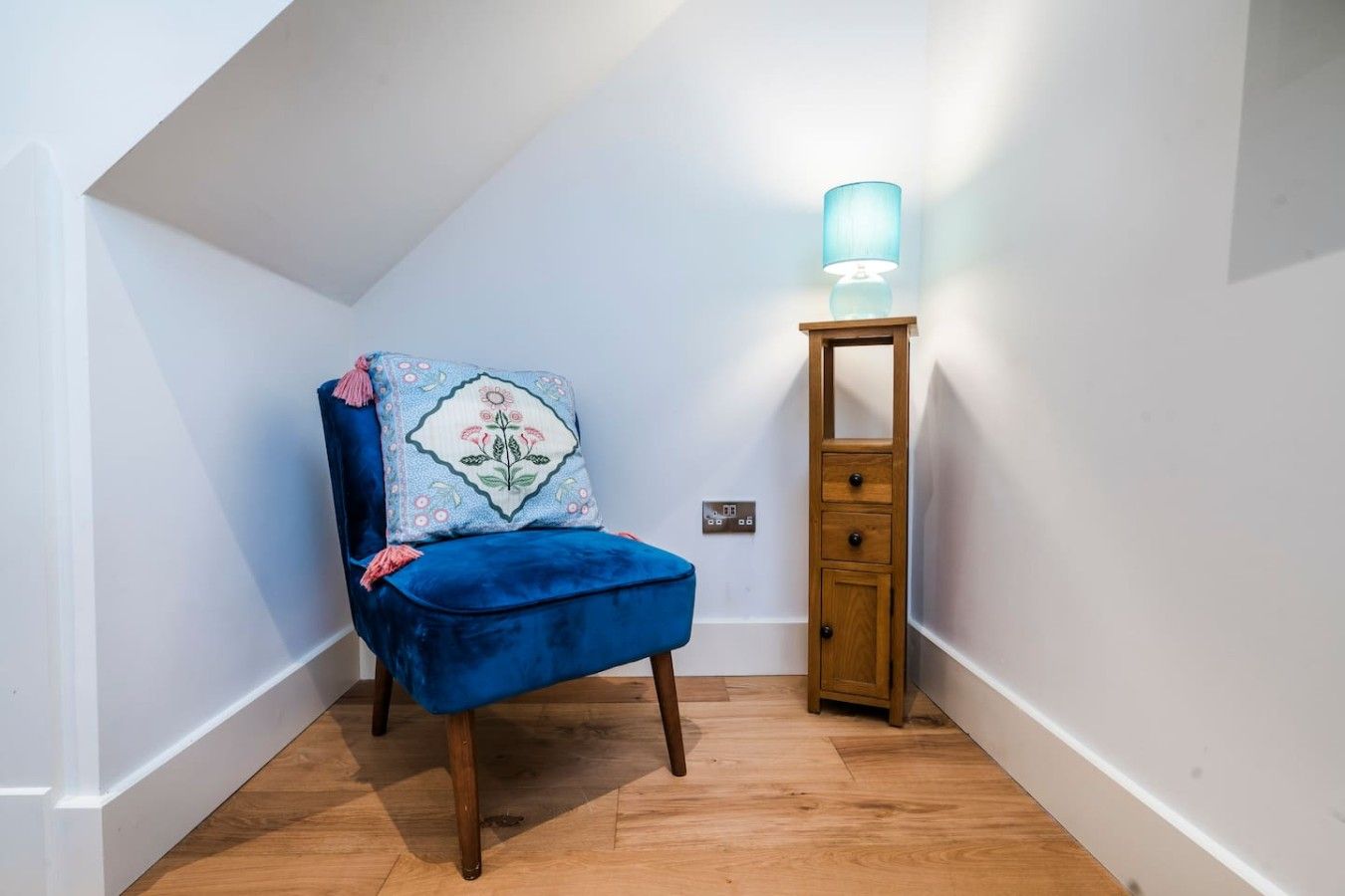 A corner room with a blue chair, a wooden cabinet, and a wall-mounted lamp.