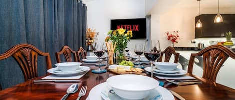 A dining area with a wooden table set for a meal, a TV mounted on the wall, and a kitchen in the background.