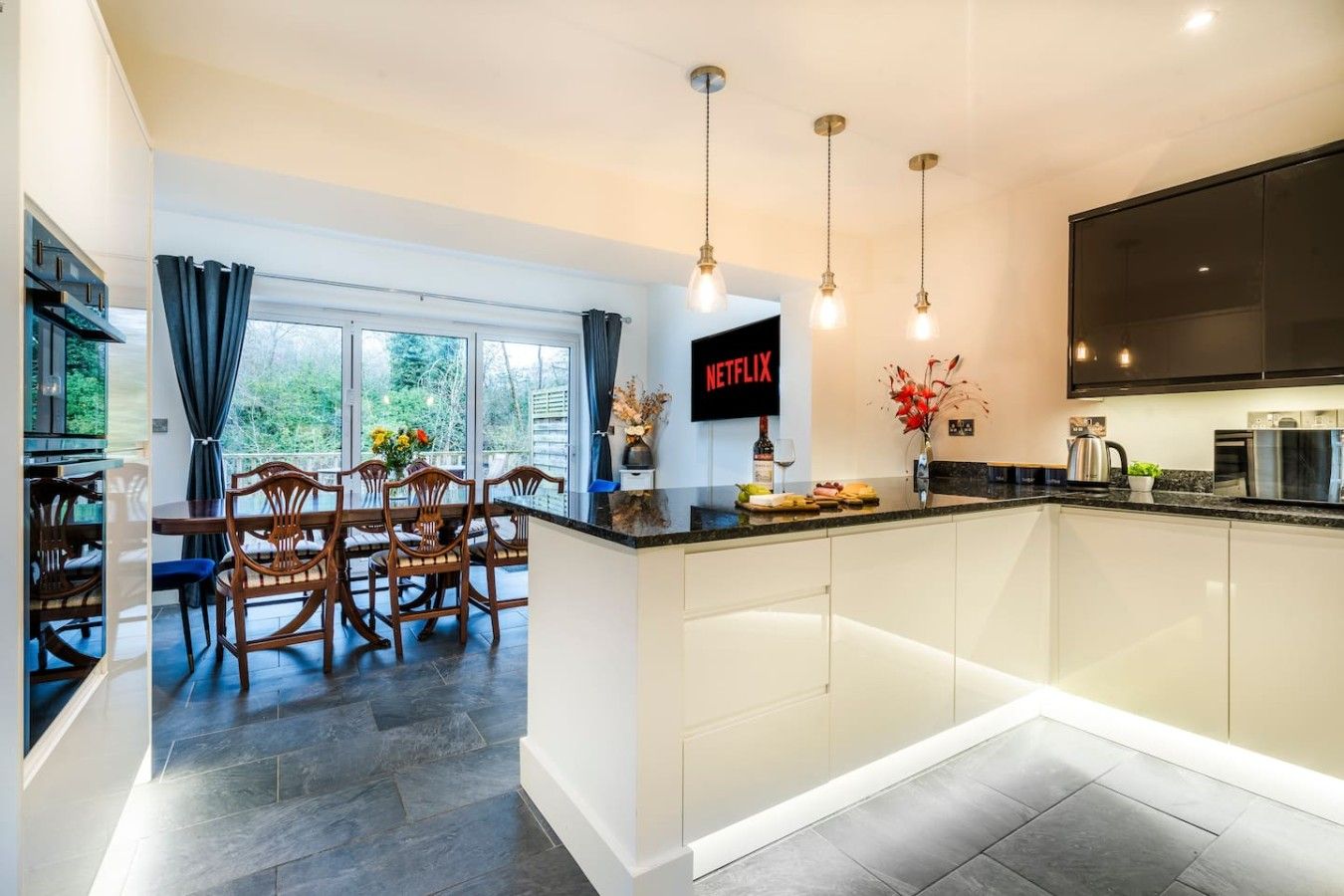 A modern kitchen with a dining area, a large TV screen, and a view of the outdoors.