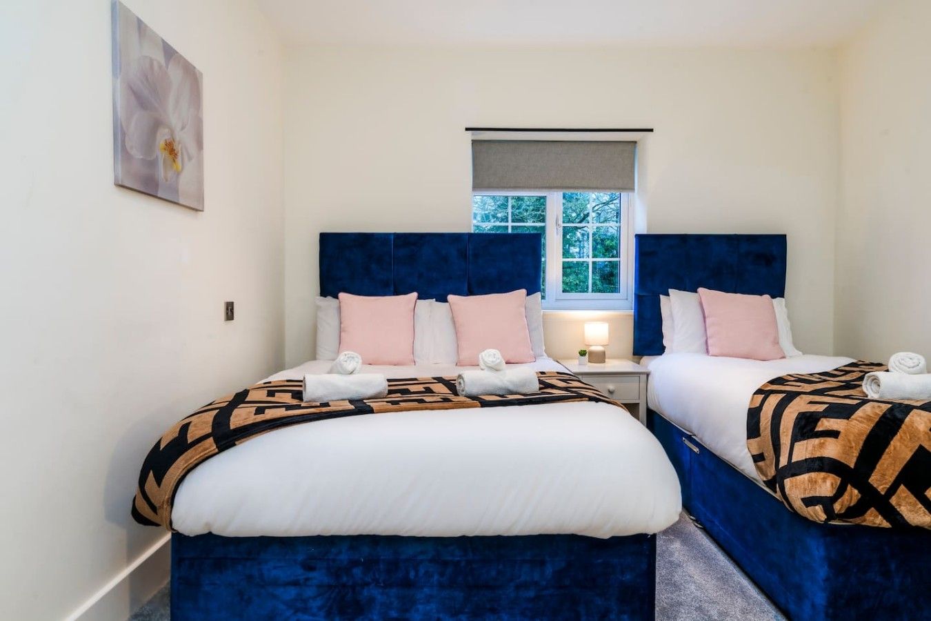 A hotel room with two beds, each with a blue headboard, white bedding, and a patterned throw blanket. There is a window with a view of trees, a bedside table with a lamp, and a painting on the wall.