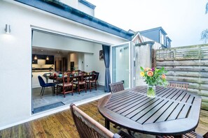 An open-plan kitchen and dining area with a wooden table and chairs, a vase with flowers, and a wooden fence in the background.