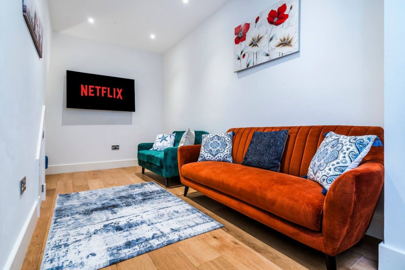 A living room with a red sofa, teal armchair, and a blue patterned rug. A flat-screen TV mounted on the wall displays 'NETFLIX'.
