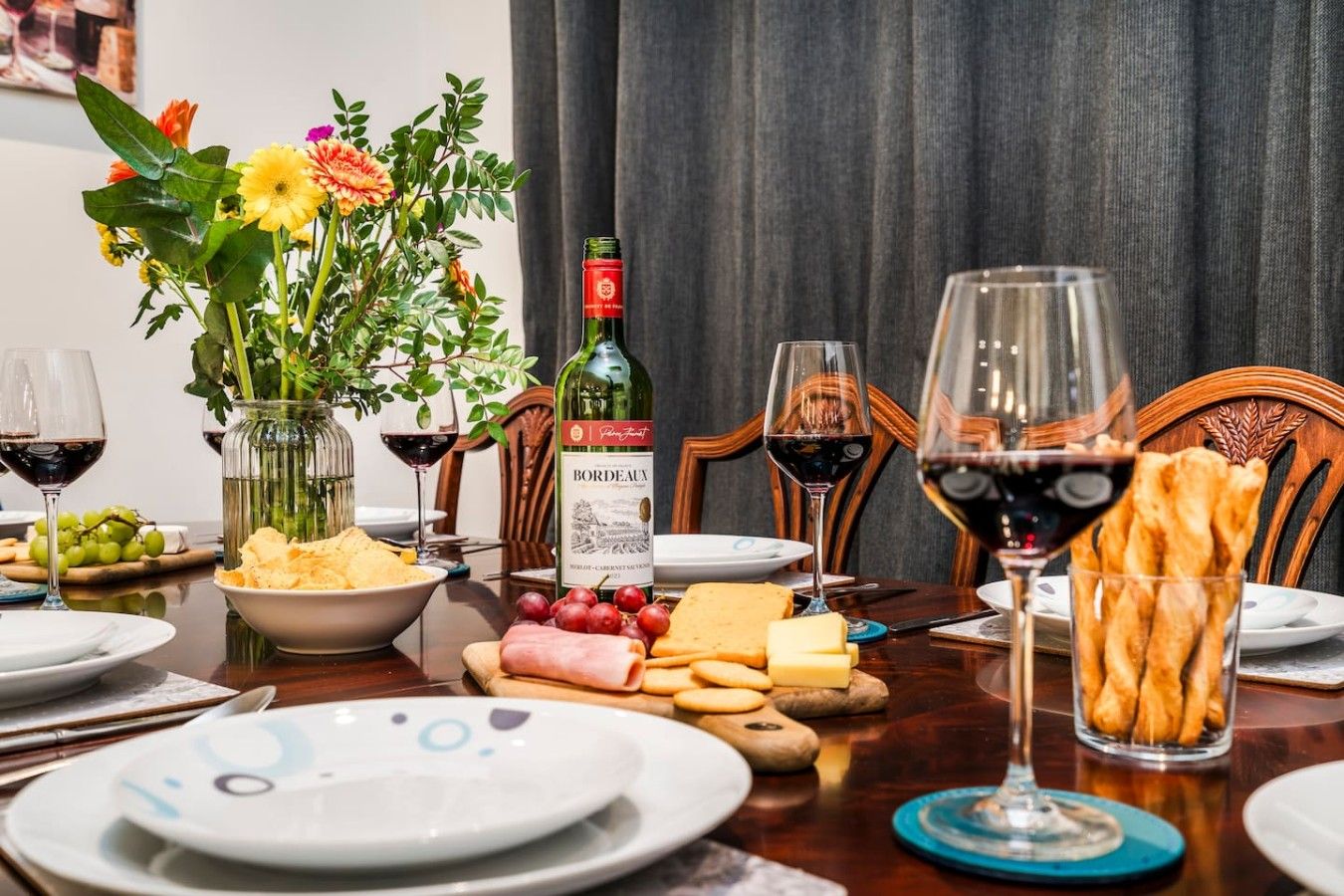 Ett matbord dukat med tallrikar, glas, en flaska Bordeauxvin, en vas med blommor och olika sorters mat.