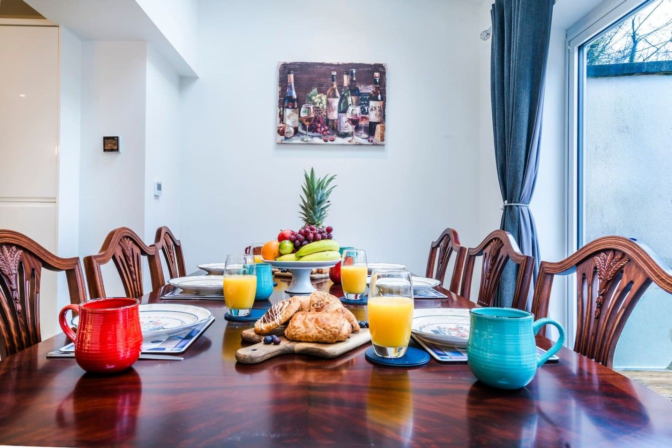Mesa posta para o café da manhã, com frutas, doces e bebidas, em um quarto com um quadro na parede e uma janela com cortinas.