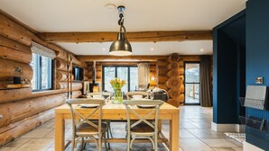 A wooden log cabin interior with a dining area featuring a wooden table and chairs, a hanging light fixture, and a view through a large window. - The Lookout (Ilkley)