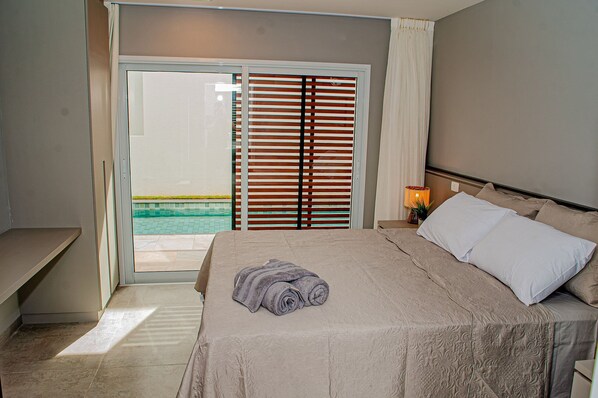 Habitación de hotel con cama, mesita de noche, lámpara y vista a una piscina a través de una puerta corredera.