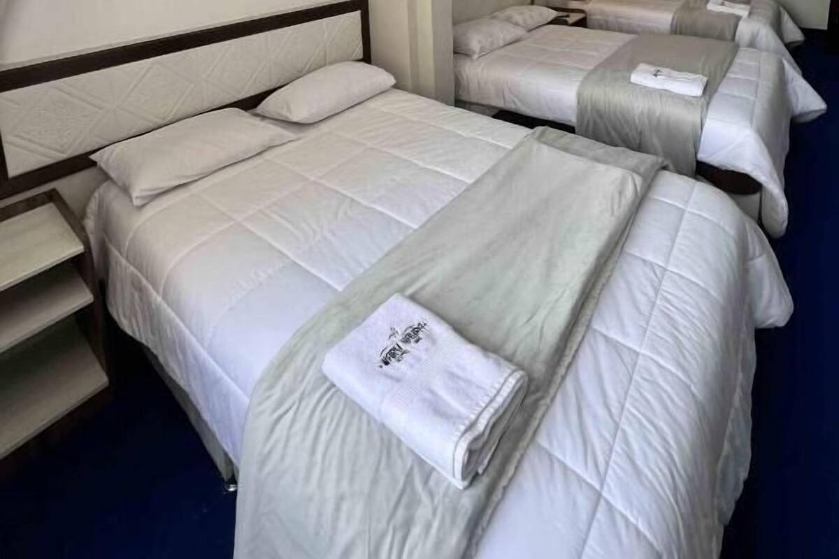 Habitación de hotel con tres camas dispuestas en fila, cada una con cabecera y mesita de noche.