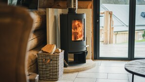 A cozy room with a wood-burning stove, a wicker basket with firewood, and a view through a glass door. - Keepers Cabin (Ilkley)