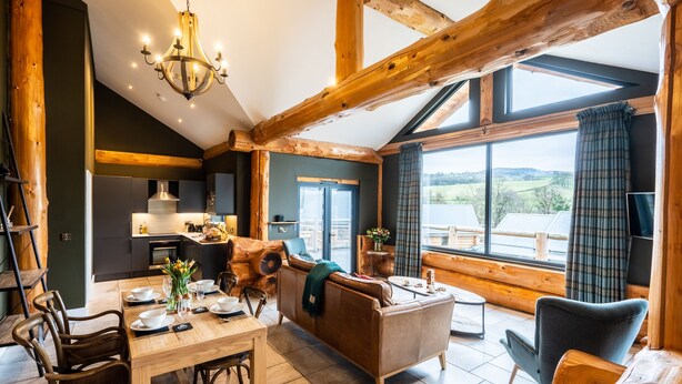 A cozy cabin interior with a dining area, sofa, and large windows offering a view of the outdoors. - Bears Den (Ilkley)