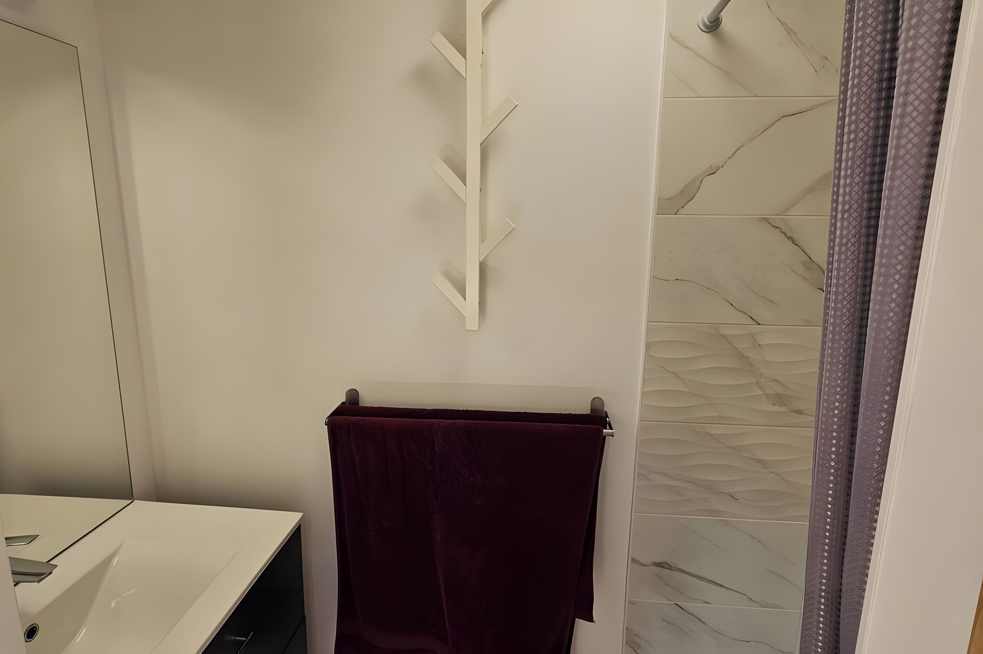 Un baño con un lavamanos blanco, un perchero con una toalla oscura y una ducha con pared de azulejos estampados.
